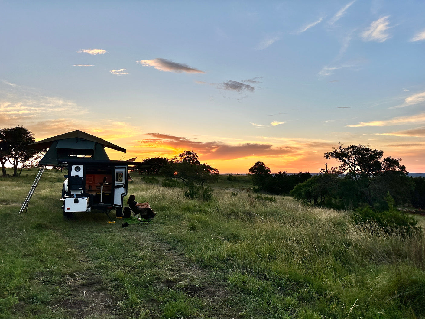 Peaceful RV rental camping at sunset. Person relaxes by a camper with a rooftop tent in a scenic, grassy landscape.