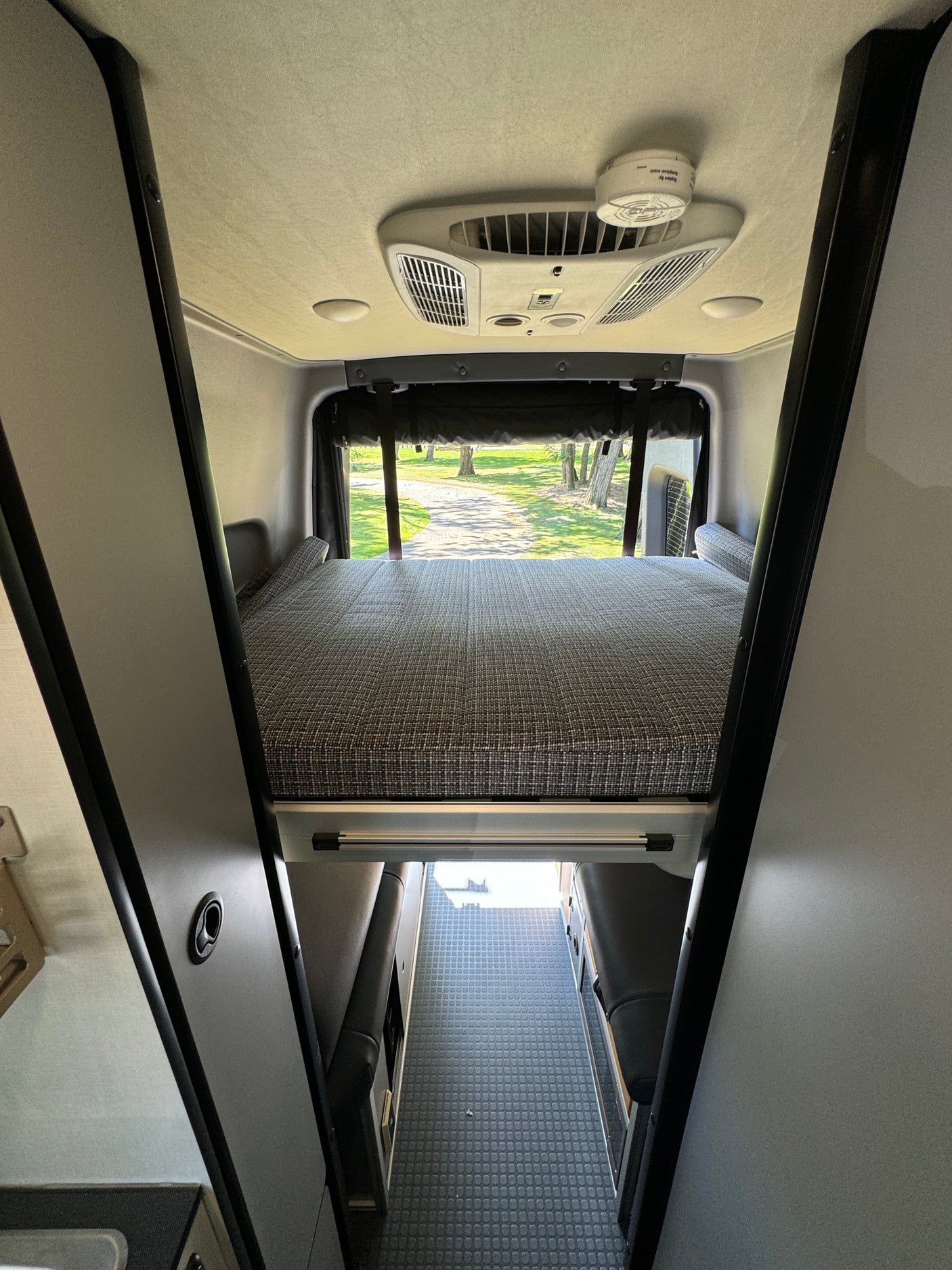 RV rental interior: A raised bed with textured mattress and outdoor view. AC unit & aisle below. Perfect for comfortable travel.