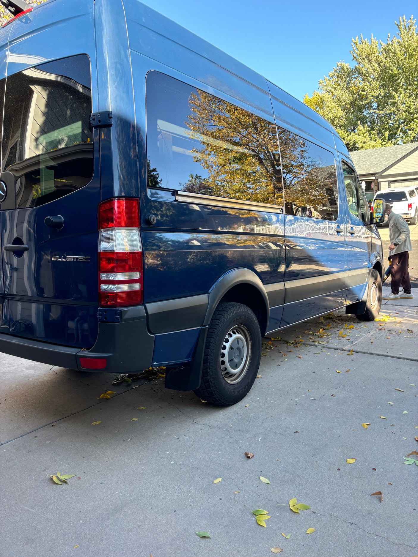 Dark blue Mercedes Sprinter van, perfect for an rv rental, parked on a driveway with autumn leaves. A person clears leaves.