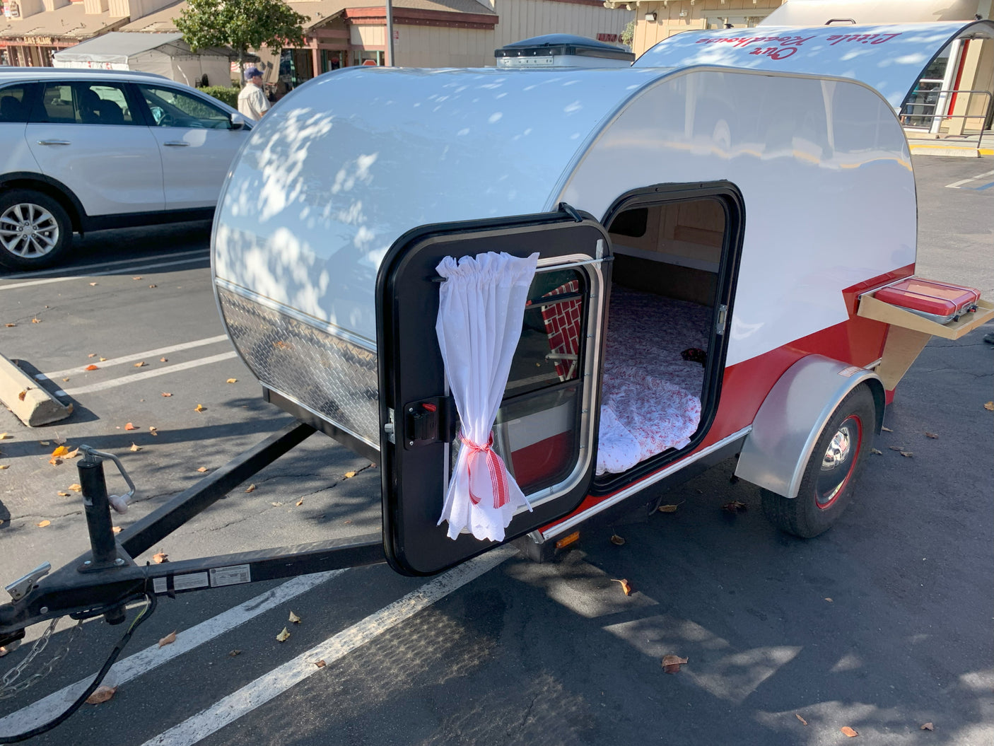 Cozy white & red teardrop camper, an ideal RV rental, with an open door showing the bed & small pull-out stove for travel.