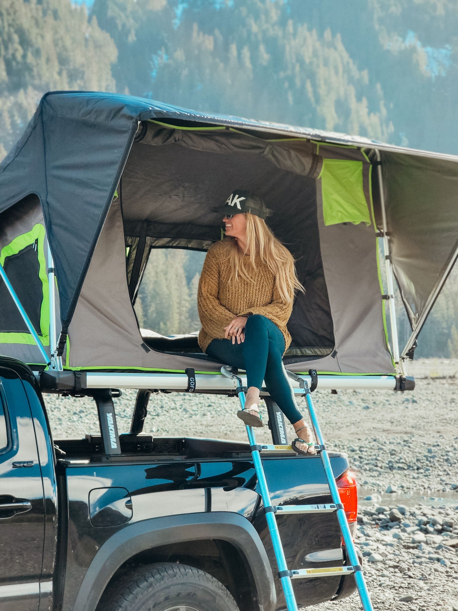 Adventure-ready RV rental! Woman relaxes in a rooftop tent atop a truck, by a scenic river.