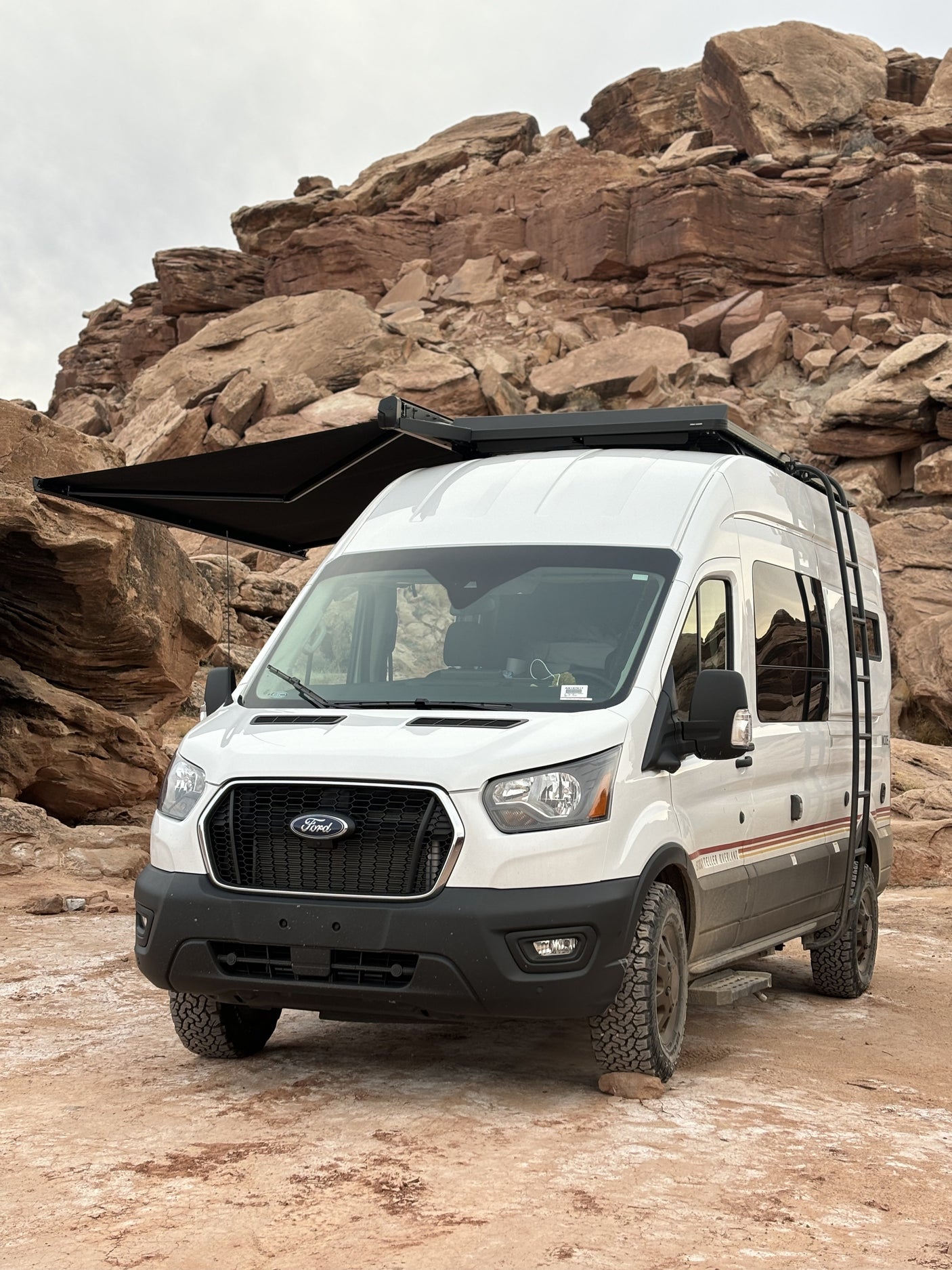 White Ford Transit camper van with extended awning, parked on rocky terrain amidst large red rock formations. Ideal RV rental for adventure.