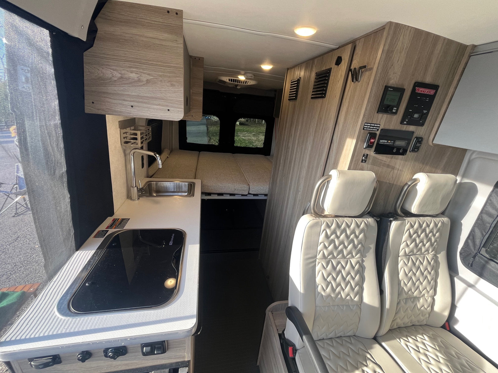 Explore this modern Winnebago rv rental interior: a full view of the kitchenette, rear bed, quilted passenger seats, and control panel wall.