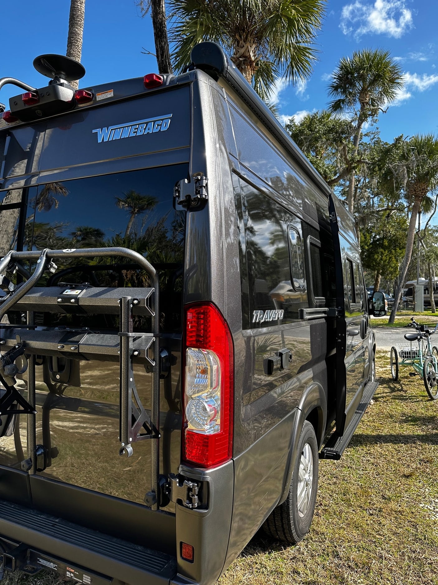 This dark Winnebago Travato RV rental is parked with a bike rack, ready for adventure under blue skies.