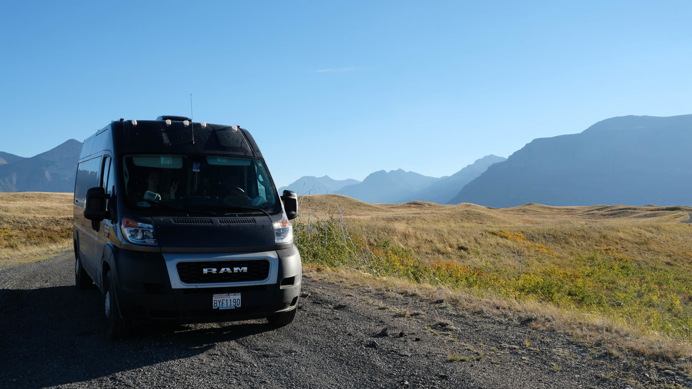 Black RAM camper van, possibly an RV rental, on a gravel road. Panoramic views of mountains & golden hills under a clear sky.