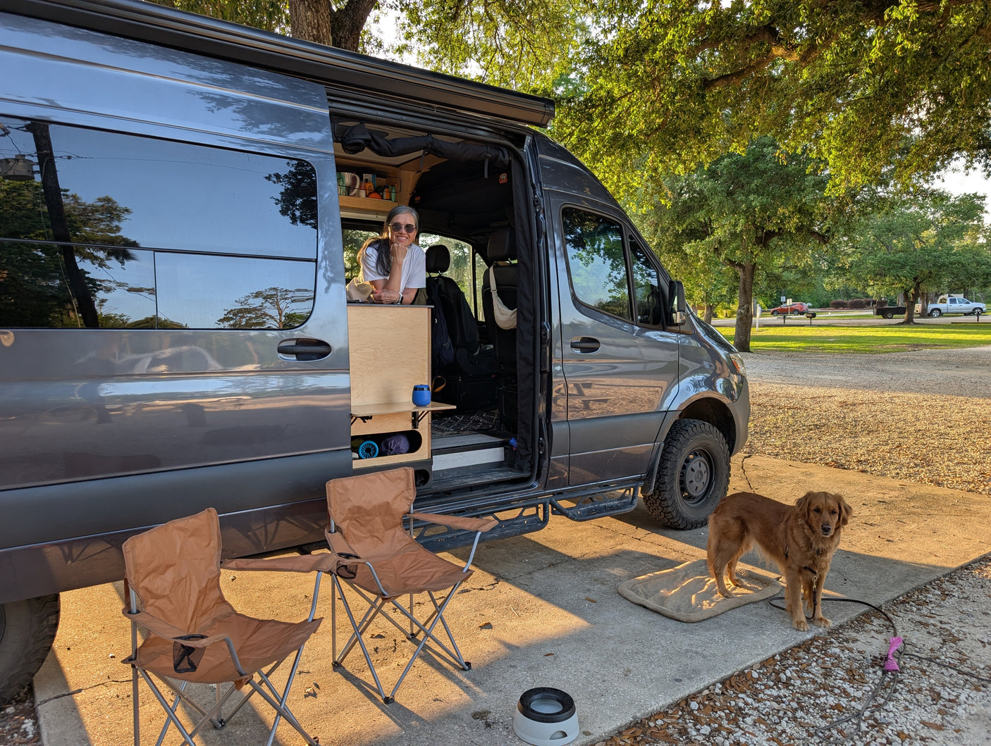 A woman and her golden retriever relax outside a gray RV rental van at a sunny campsite, with folding chairs. Perfect for #vanlife.