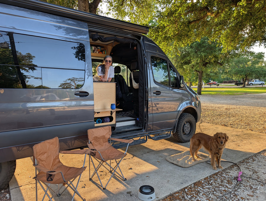 A woman and her golden retriever relax outside a gray RV rental van at a sunny campsite, with folding chairs. Perfect for #vanlife.