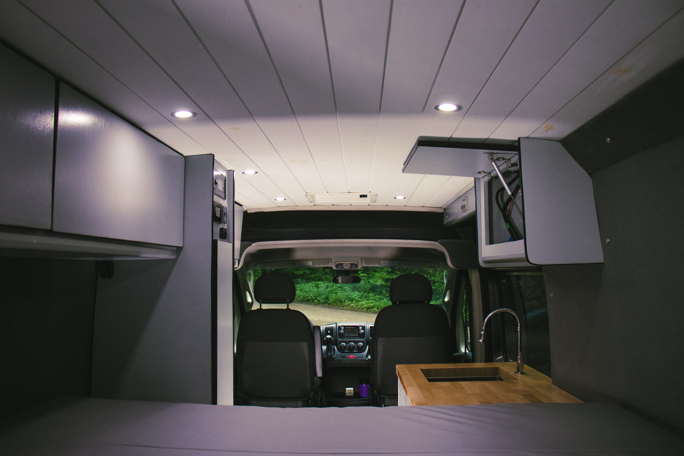 Modern RV rental interior: featuring a cozy bed, sleek kitchen, and ample overhead storage, looking towards the driver's cabin. — photo 7