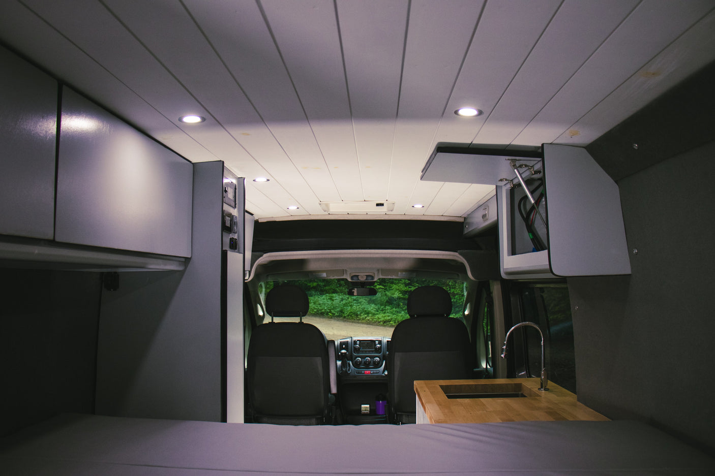 Modern RV rental interior: featuring a cozy bed, sleek kitchen, and ample overhead storage, looking towards the driver's cabin.