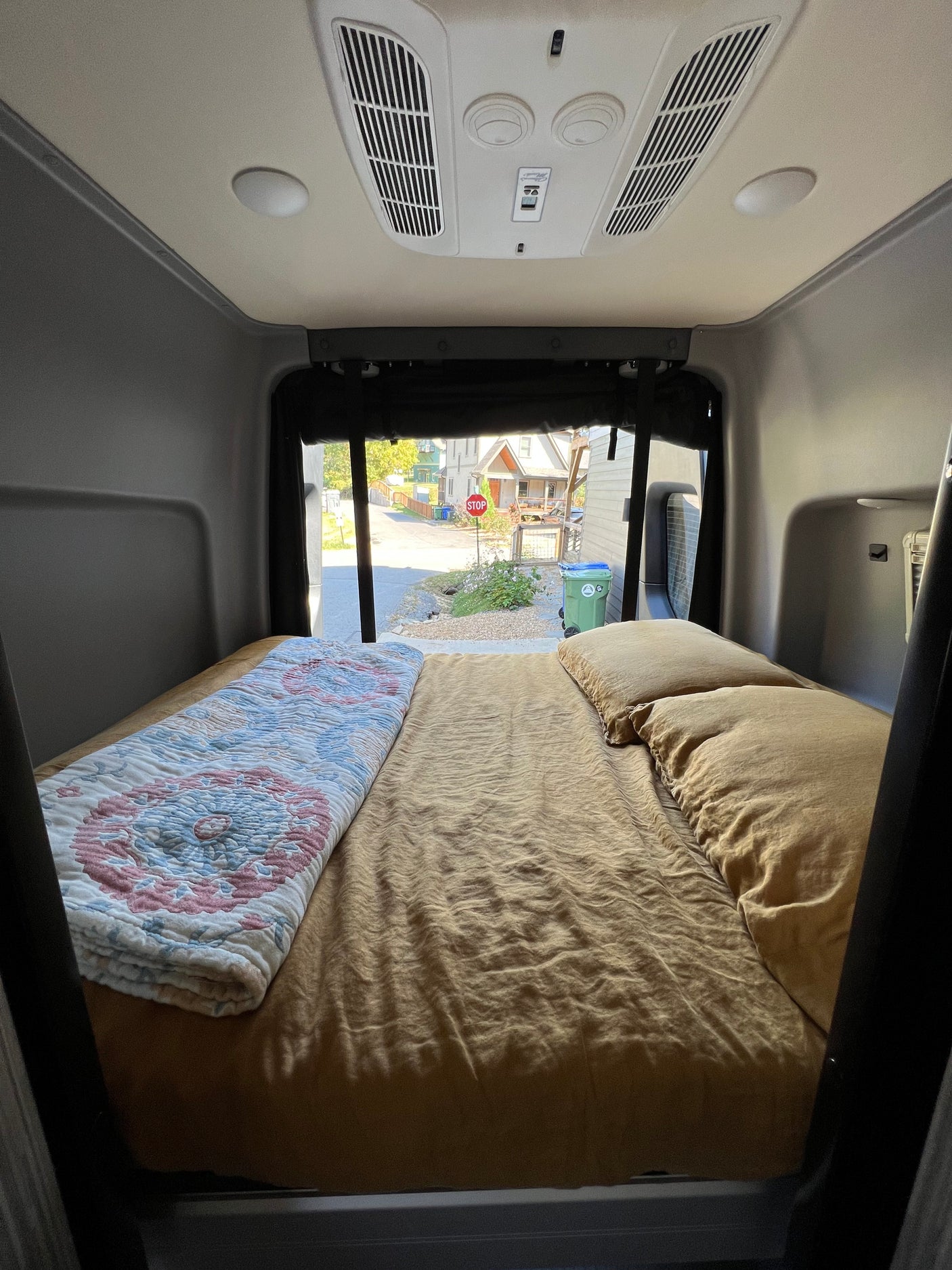 RV rental interior with a cozy bed, AC, and a view of a residential street from the back of the camper van.