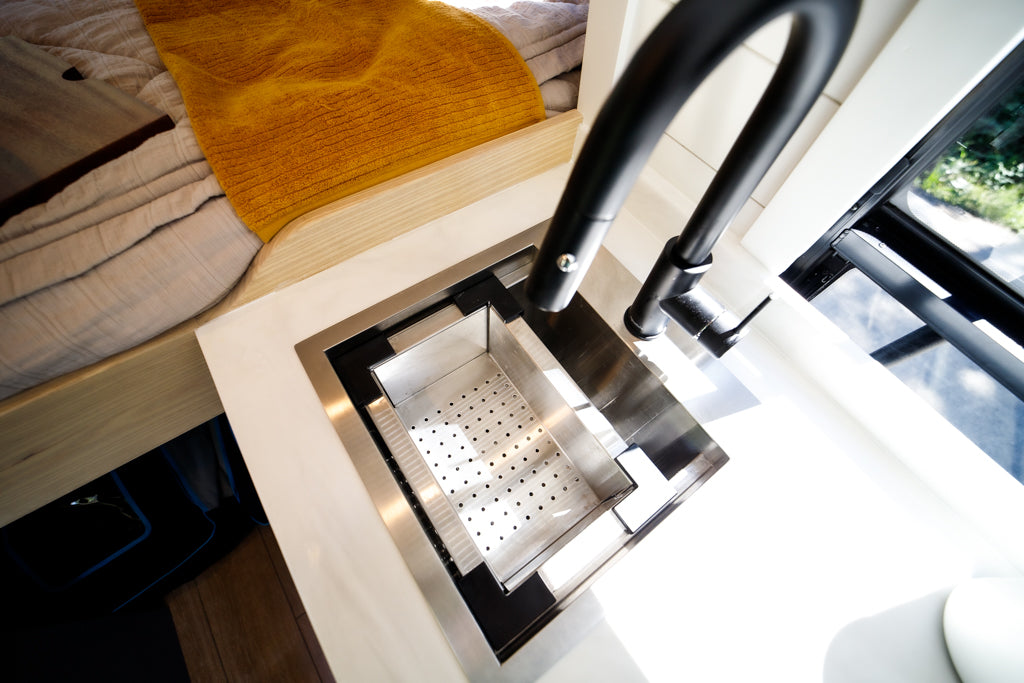 Modern RV rental interior with a sleek kitchen sink, colander insert, and black faucet next to a cozy bed setup.