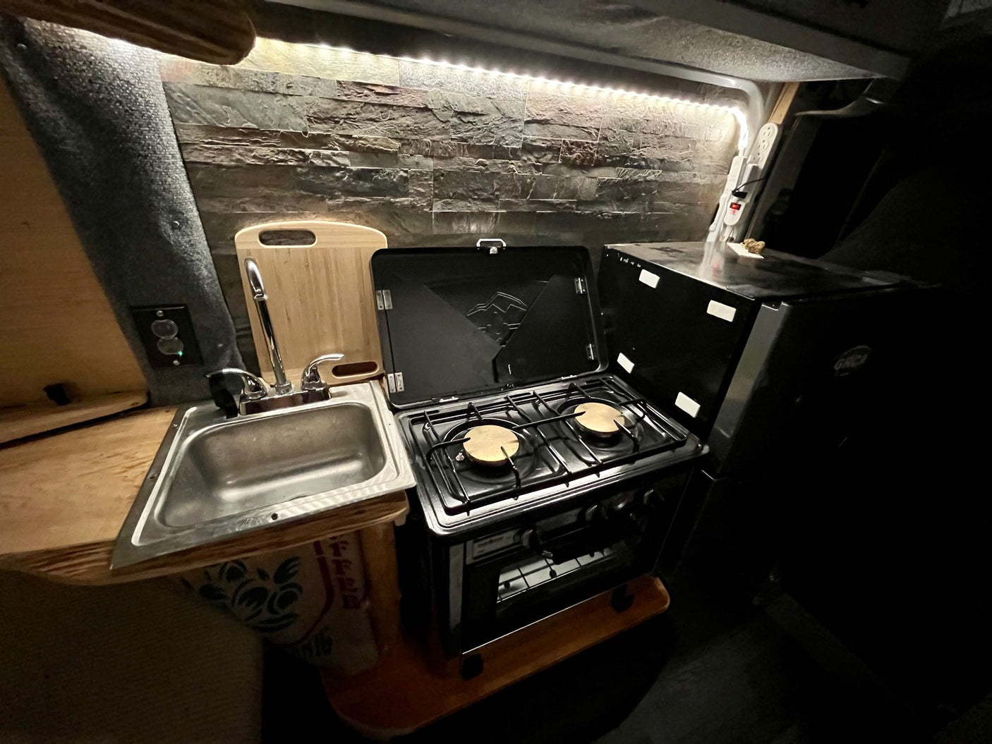 This RV rental's compact kitchen boasts a sink, two-burner stove with oven, and a fridge, all illuminated by LED strips for comfortable mobile living.