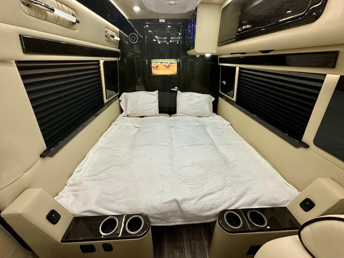 Interior of a luxury RV rental featuring a large bed with white bedding, TV, built-in cup holders, and pleated window blinds.