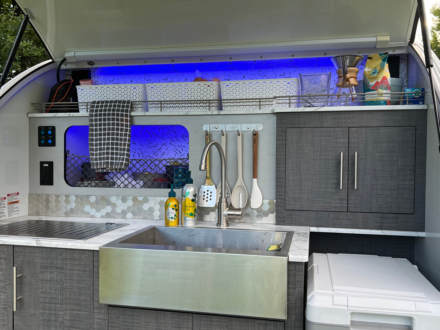 This RV rental's sleek outdoor kitchen includes a sink, Dometic stove, and ample, illuminated storage with blue accent lighting.
