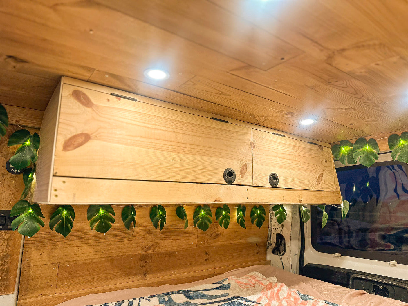Overhead wooden cabinets with leaf string lights, a bed, and ceiling lights inside a cozy RV rental van.