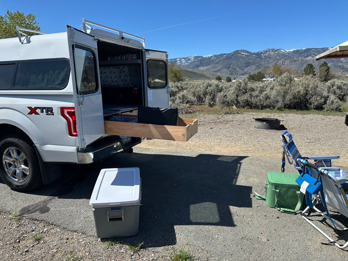Scenic rv rental setup: a truck camper with open rear doors & pull-out storage, chairs, coolers, and mountains.