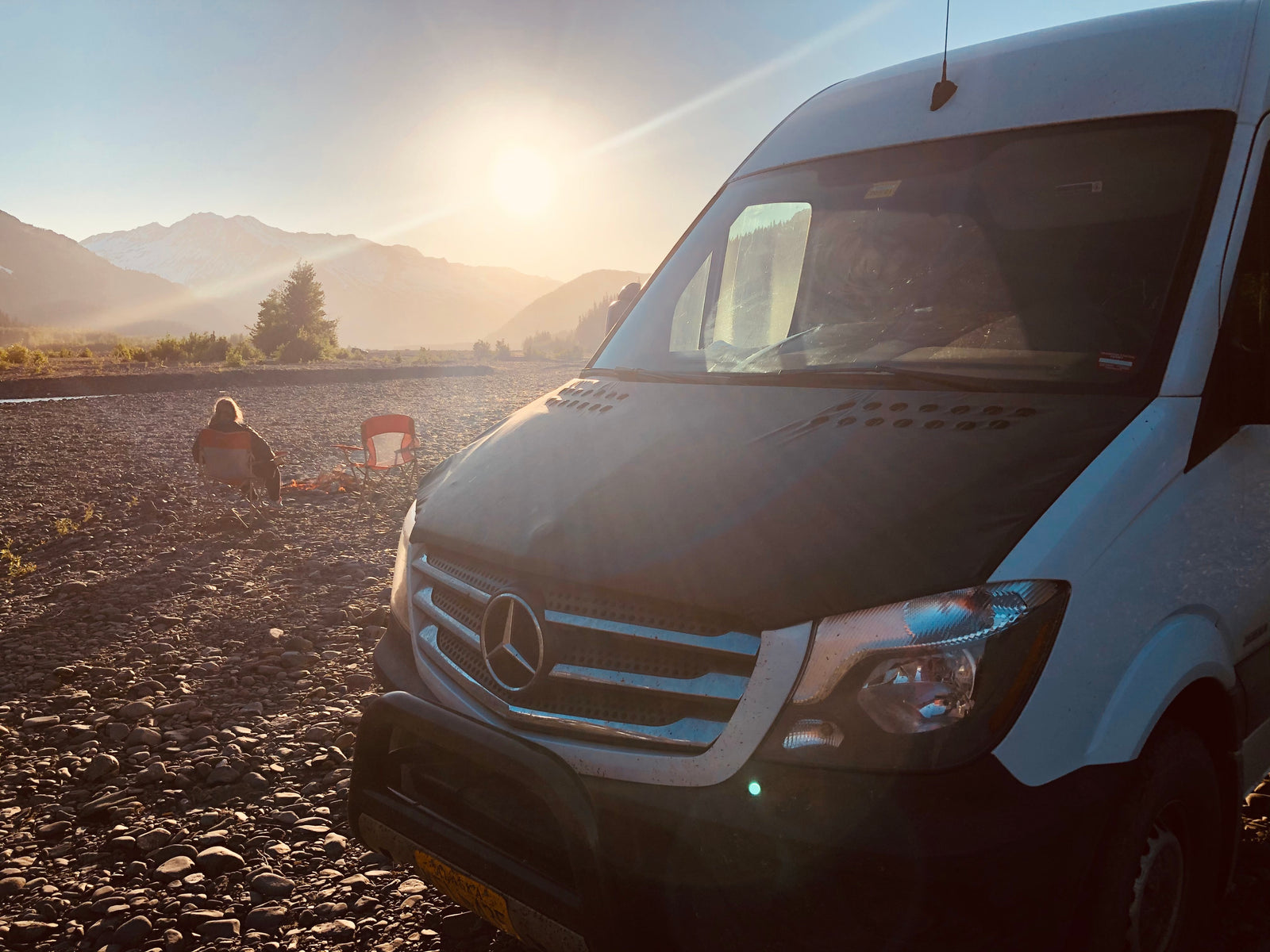 Adventure awaits! An RV rental Sprinter van parked on a rocky riverbed at sunset, with mountains & a person camping.