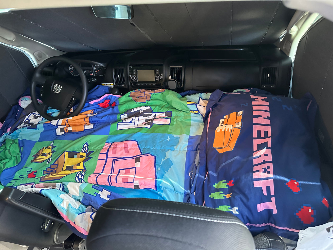 Cozy RV rental setup: Minecraft bedding fills the van's front for sleeping. Dashboard & sunshade visible.