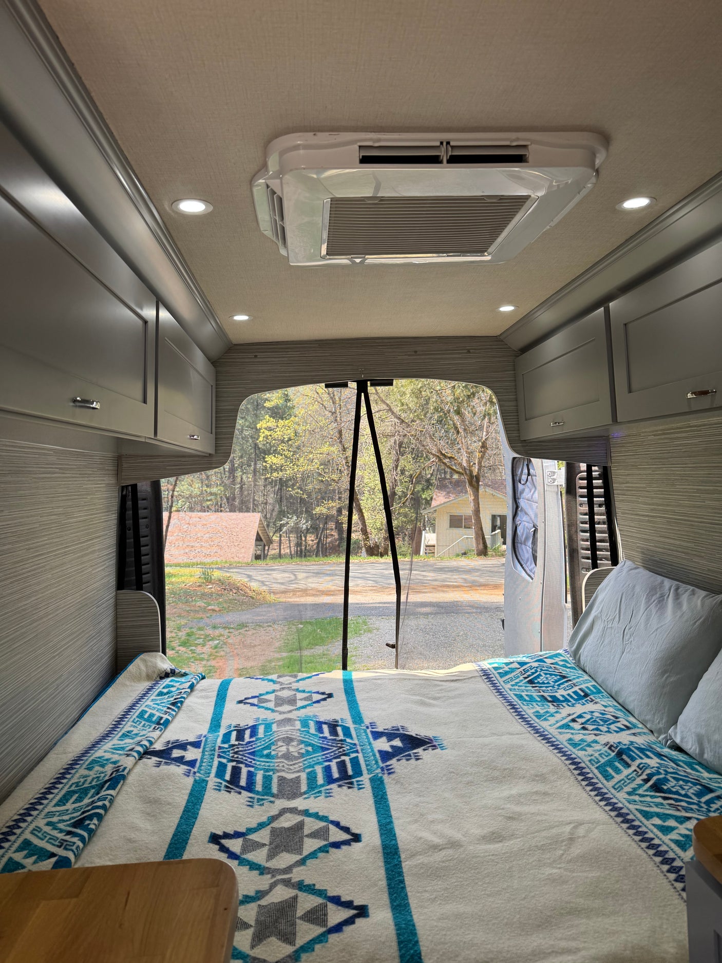 RV rental interior: A cozy bed with a blue patterned blanket, overhead cabinets, and open back doors reveal a wooded outdoor scene.