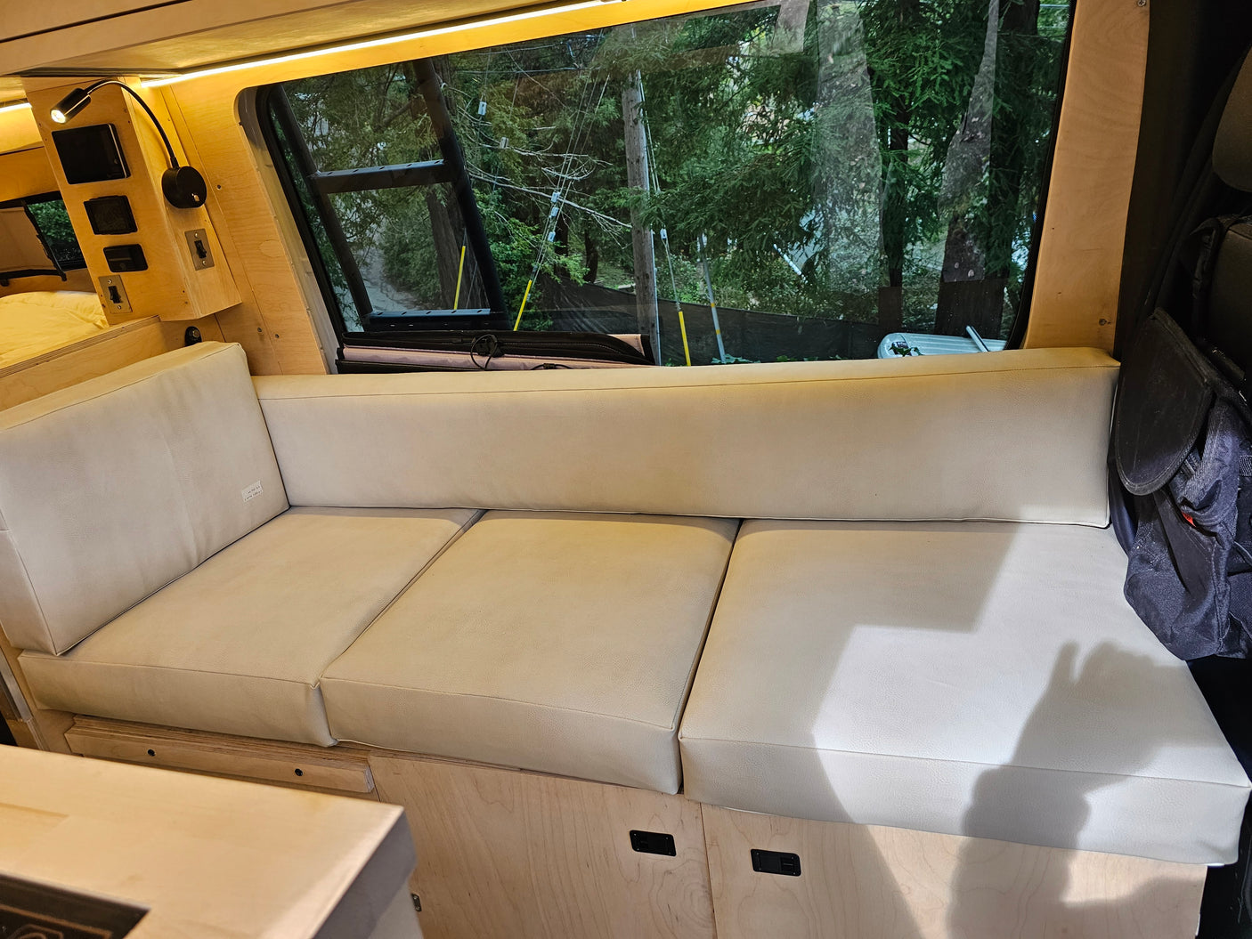 Inside an RV rental: light wood interior with a long beige bench seat, storage, and a large window showcasing outside trees.