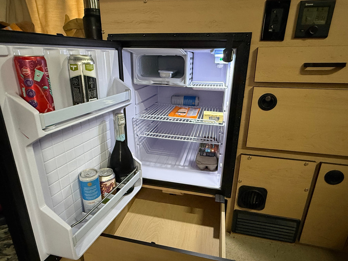An open rv rental fridge stocked with cans, champagne, butter, eggs, and a drawer pulled out below.