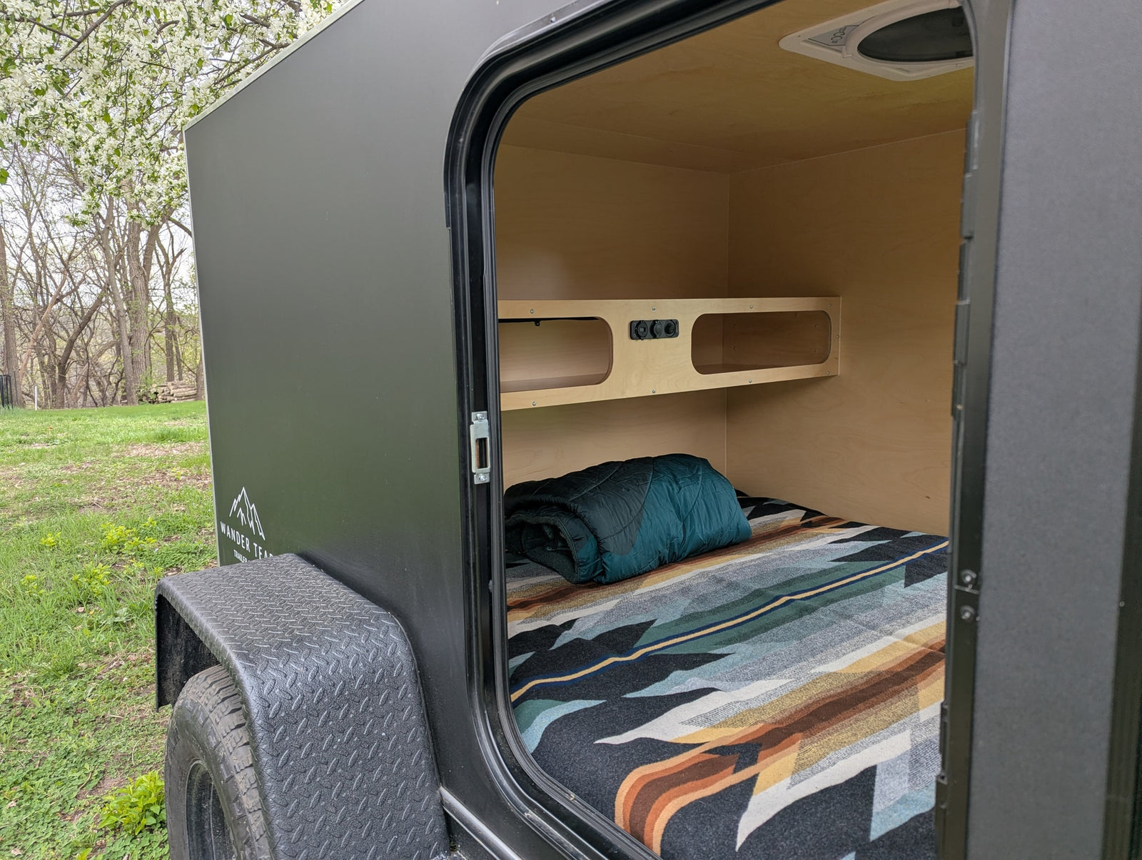 RV rental teardrop camper interior: cozy bed, shelf with power outlets, and ceiling vent, set in a blooming natural scene.