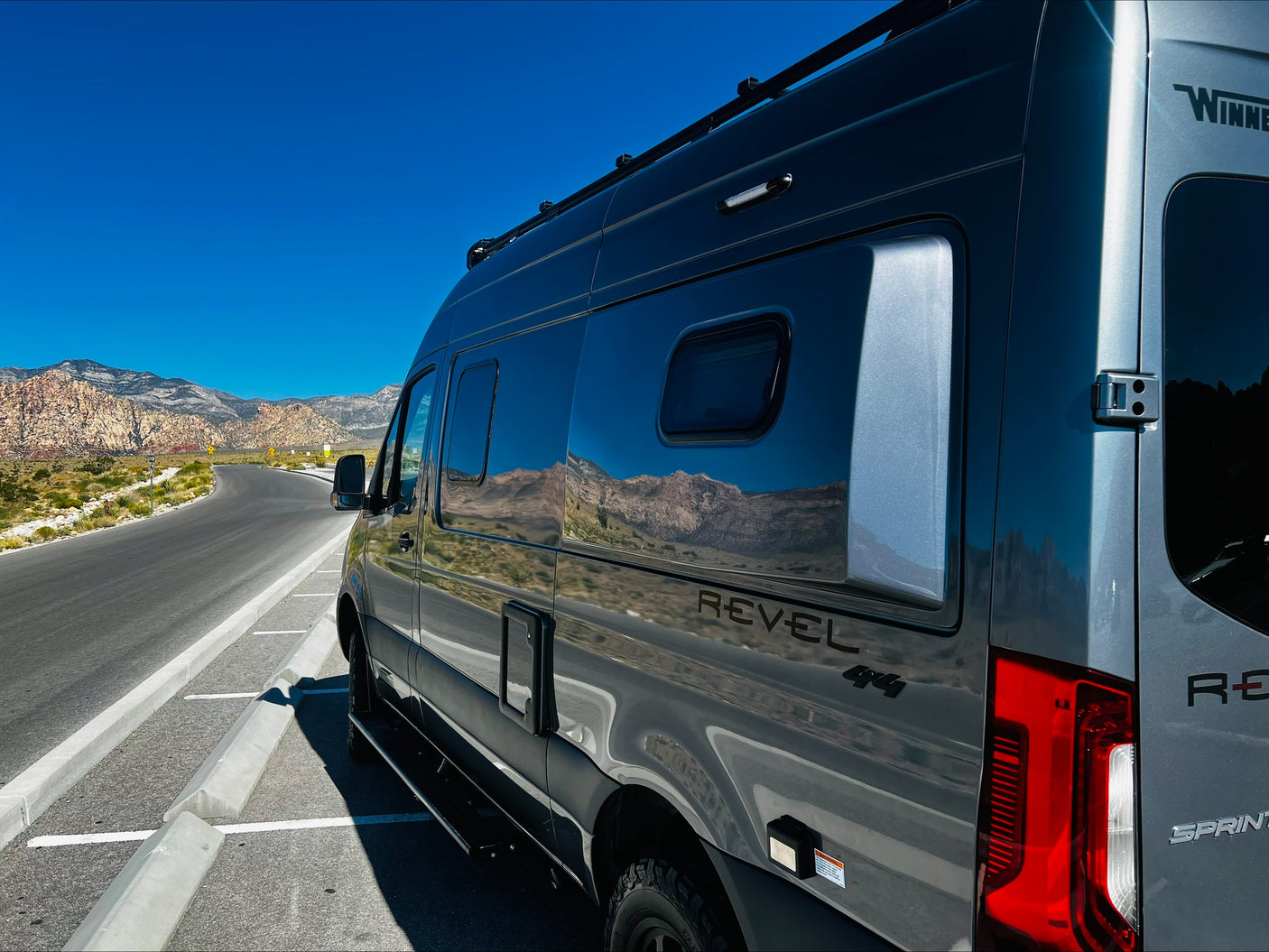 Gray Winnebago Revel 4x4 RV rental parked on a scenic desert mountain road, reflecting the blue sky and mountains.