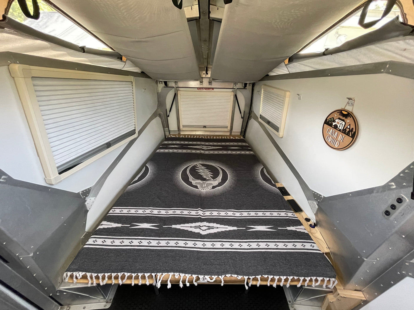 Inside a cozy pop-top RV rental, featuring a bed with a Grateful Dead blanket, window blinds, and an EXIT/SORTIE sign.