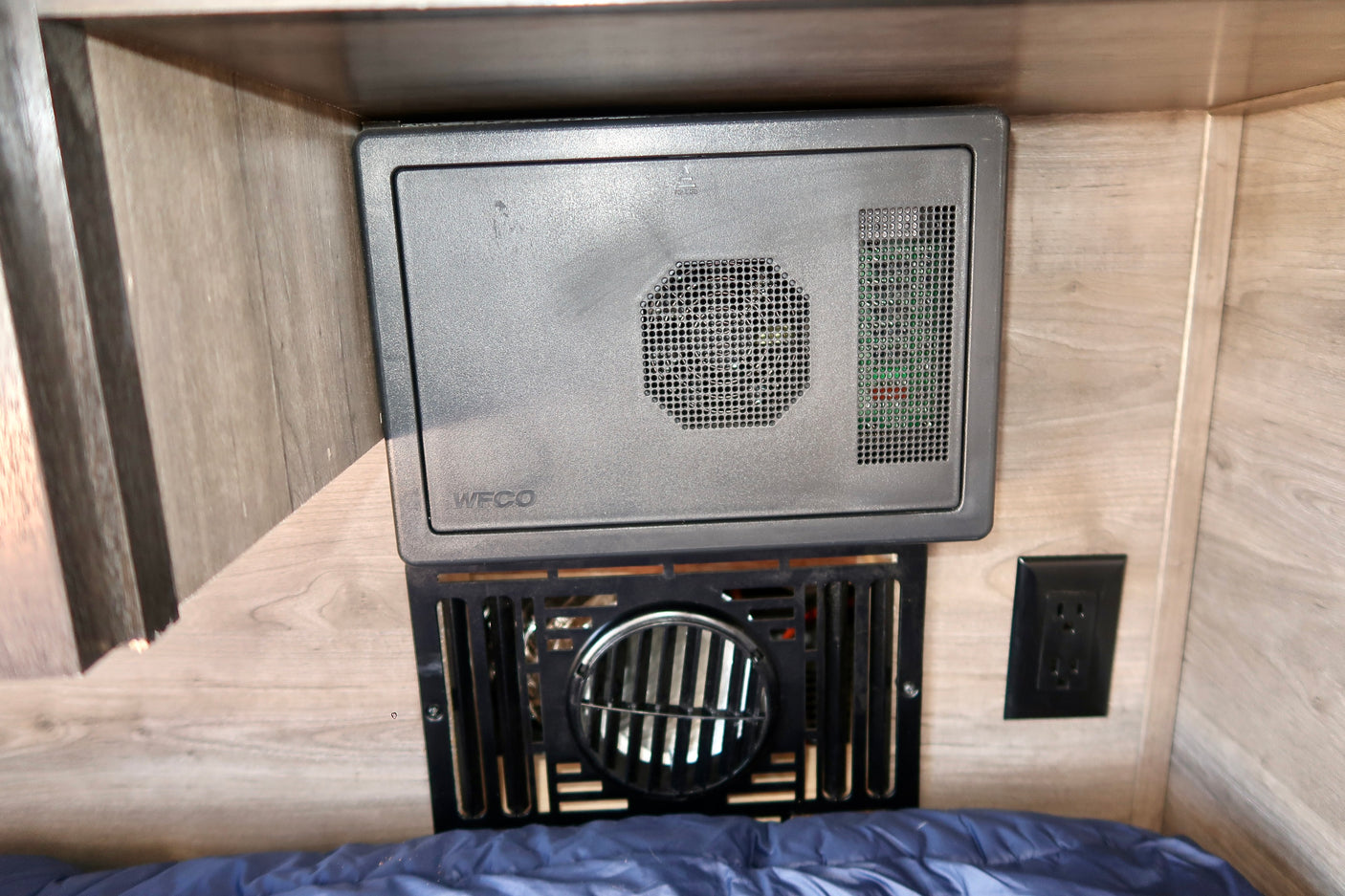 RV rental interior features: WFCO power converter/fuse box, furnace vent, and wall outlet mounted above a bed.