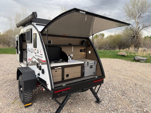 This RV rental is a white Radical Bushwacker teardrop trailer with its rear kitchen galley open, revealing a stove and cooler for outdoor camping.
