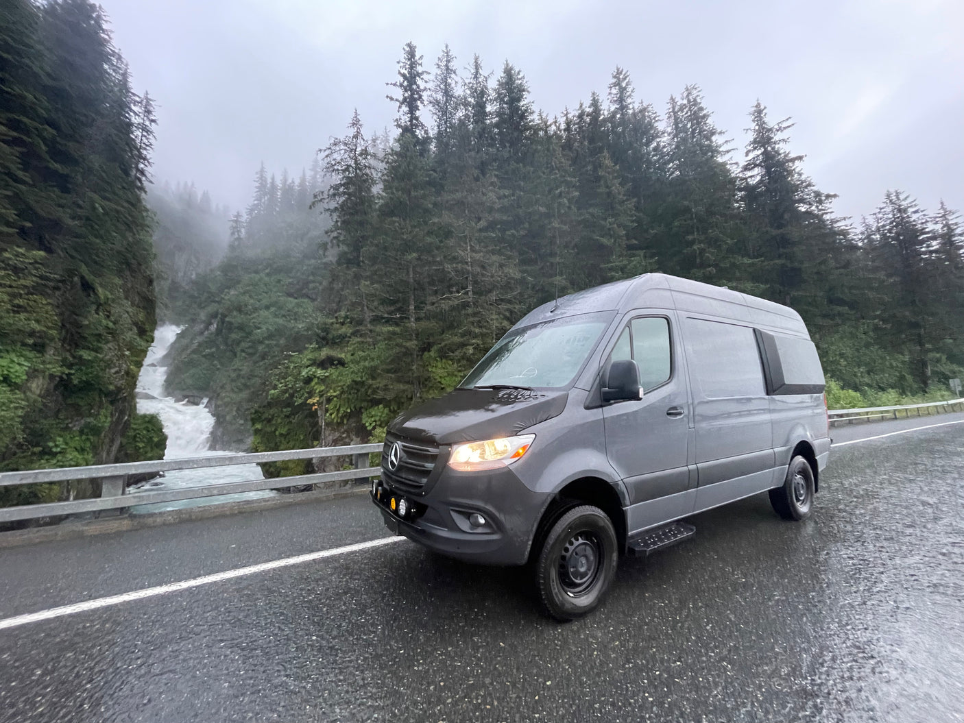 Grey Sprinter RV rental on a wet road beside a majestic waterfall and misty forest. Ideal for scenic adventures.