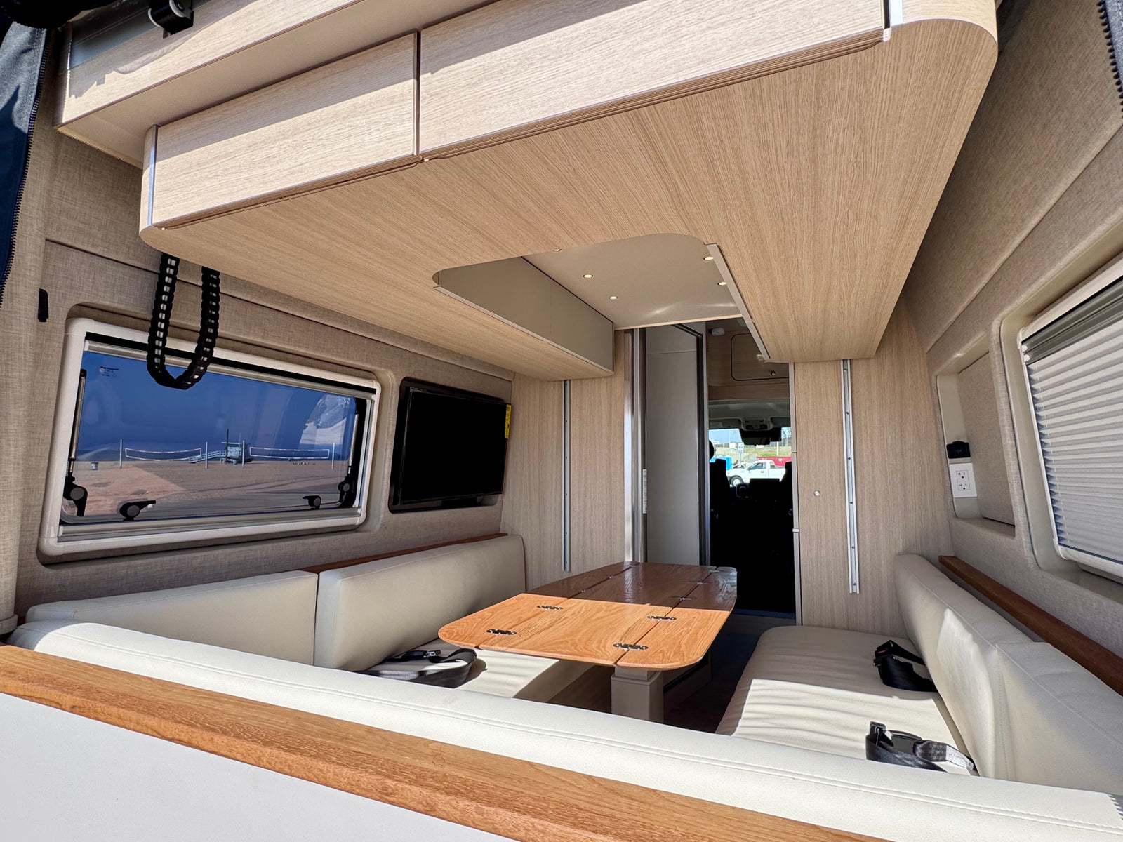 Modern RV rental interior with light wood finishes, a dining table, comfortable benches, and a clear view of a beach.