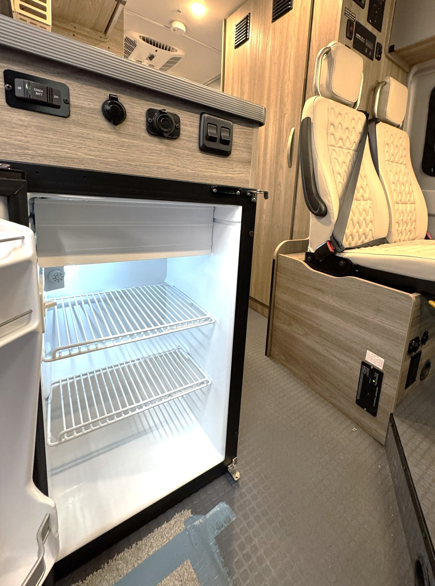 RV rental interior featuring an open, empty refrigerator, two quilted passenger seats, and a control panel with power outlets and switches.