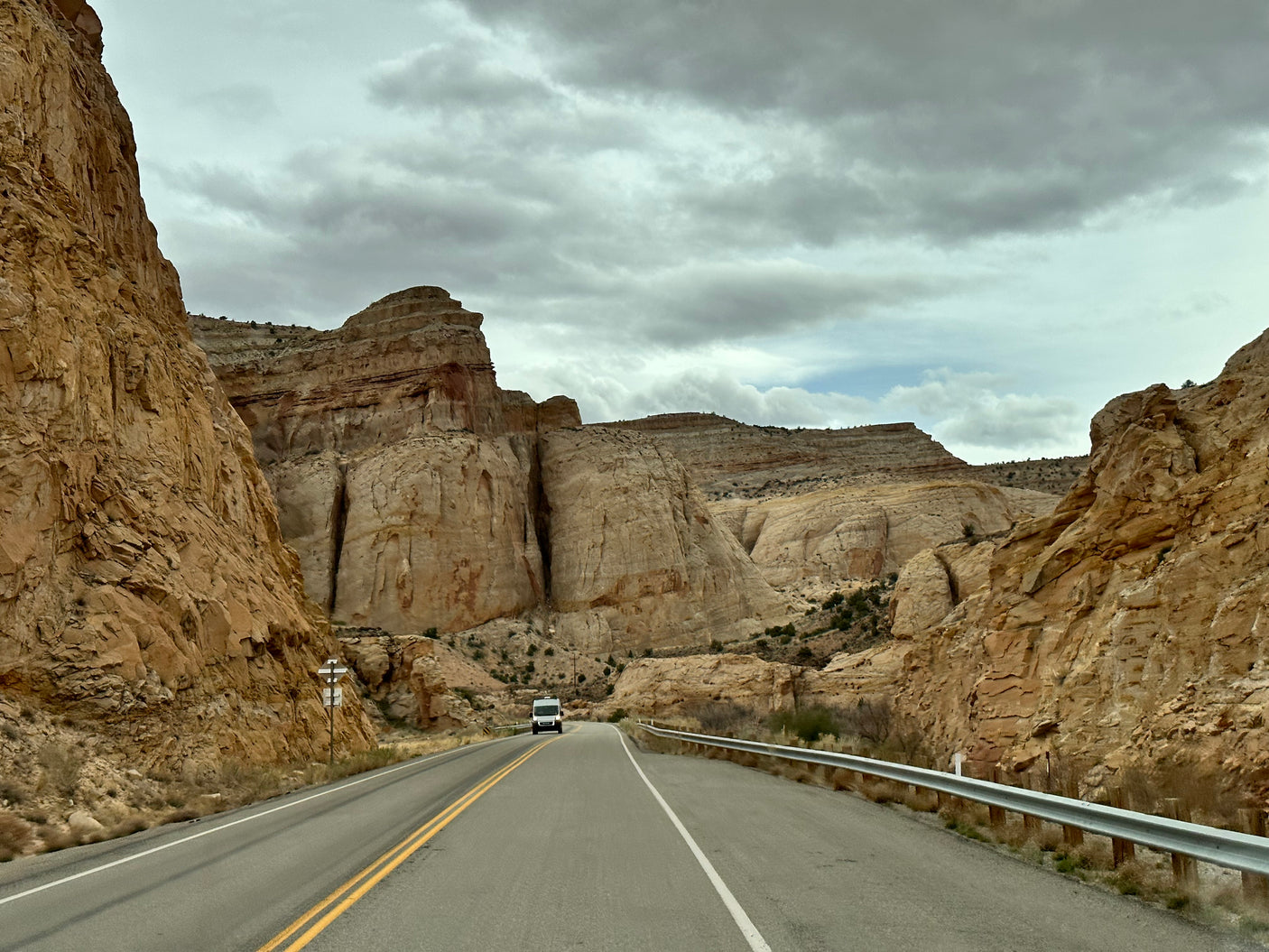 An RV rental van travels a road winding through towering canyon rock formations under a cloudy sky.