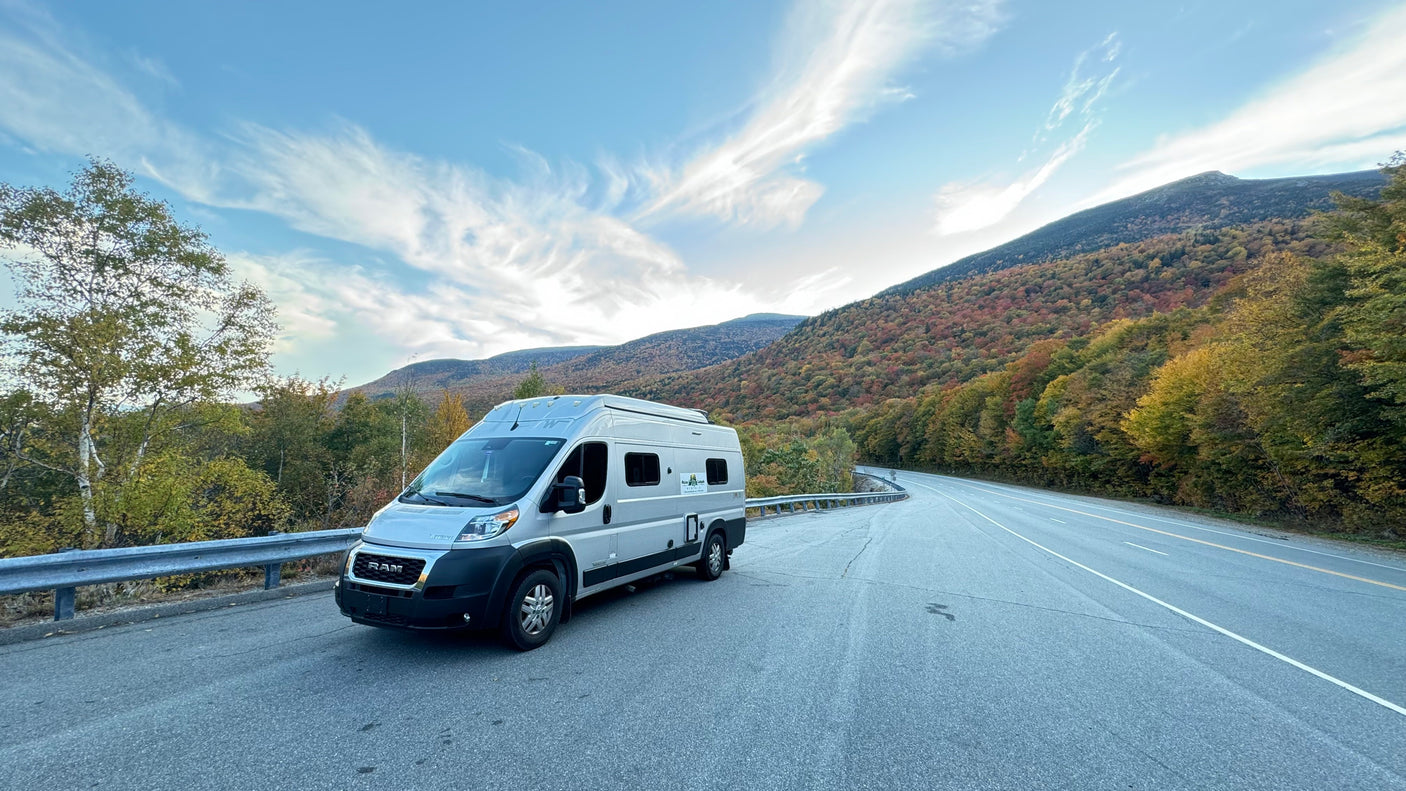 A silver RV rental is parked on a scenic mountain road, surrounded by vibrant autumn trees under a blue sky.