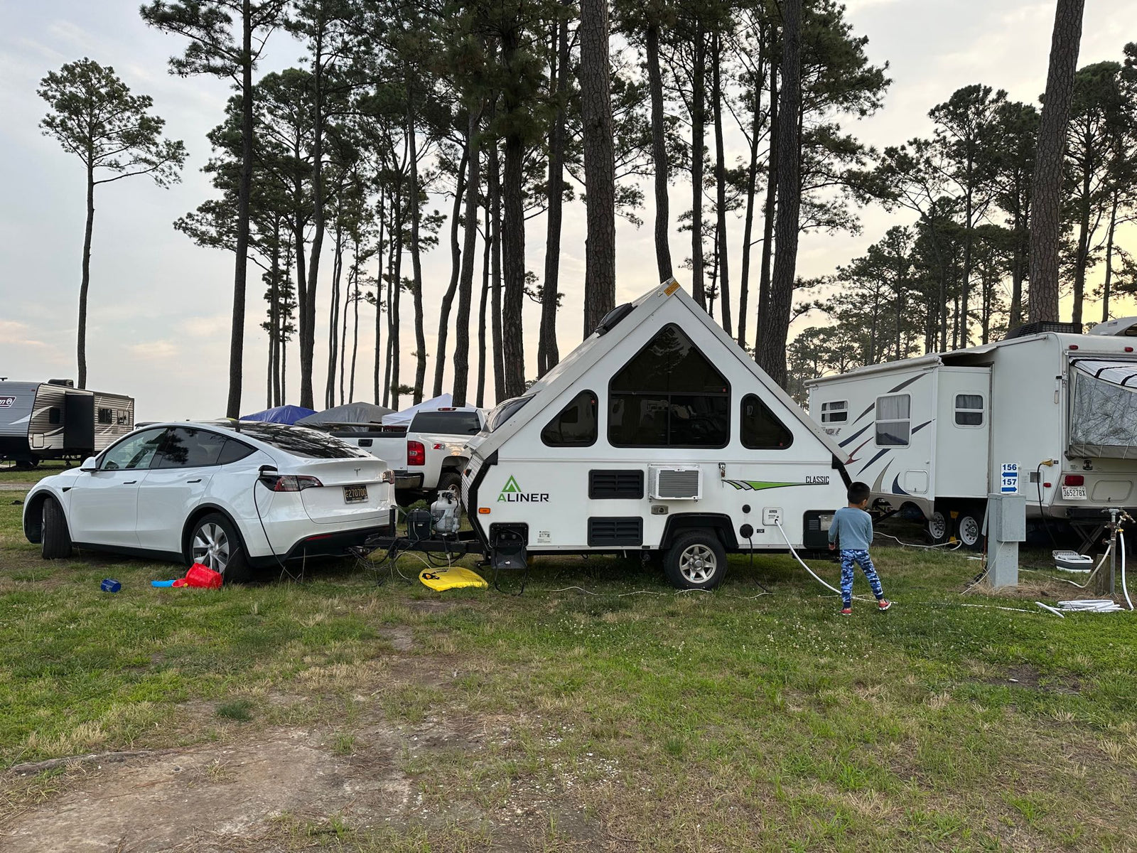 Electric vehicle charging an RV rental at a scenic campground. A child stands by the A-Liner travel trailer