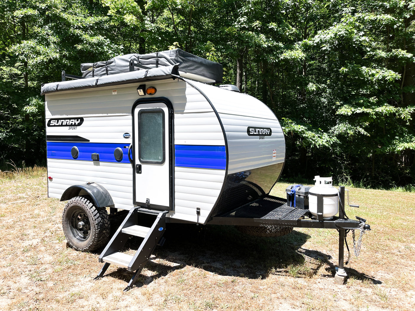 A Sunray Sport compact RV rental, a white and blue camper trailer, parked on dry ground with green trees behind it.