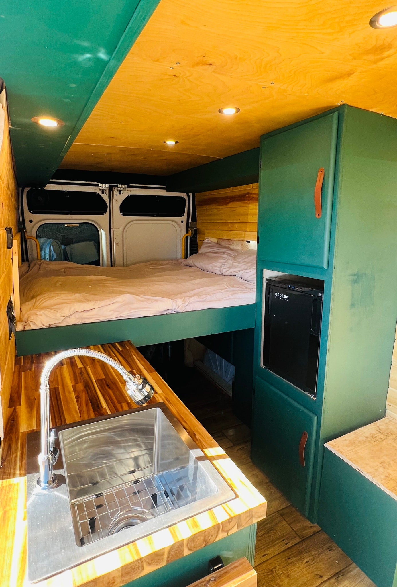 RV rental: Cozy high bed, sink kitchen, fridge, and green cabinets in a rustic wood-finished camper van interior.