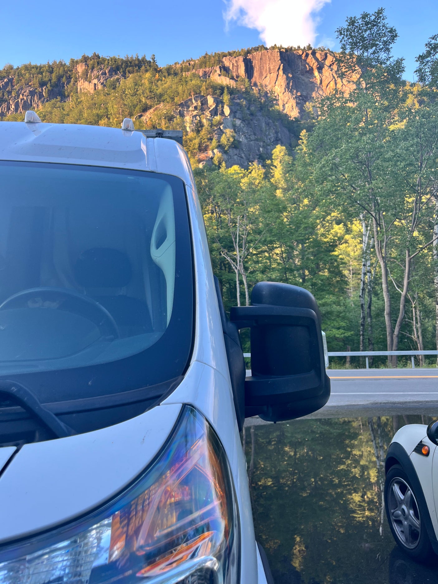 White RV rental parked by water reflecting trees & a small car. Rocky mountains & lush forest fill the background.