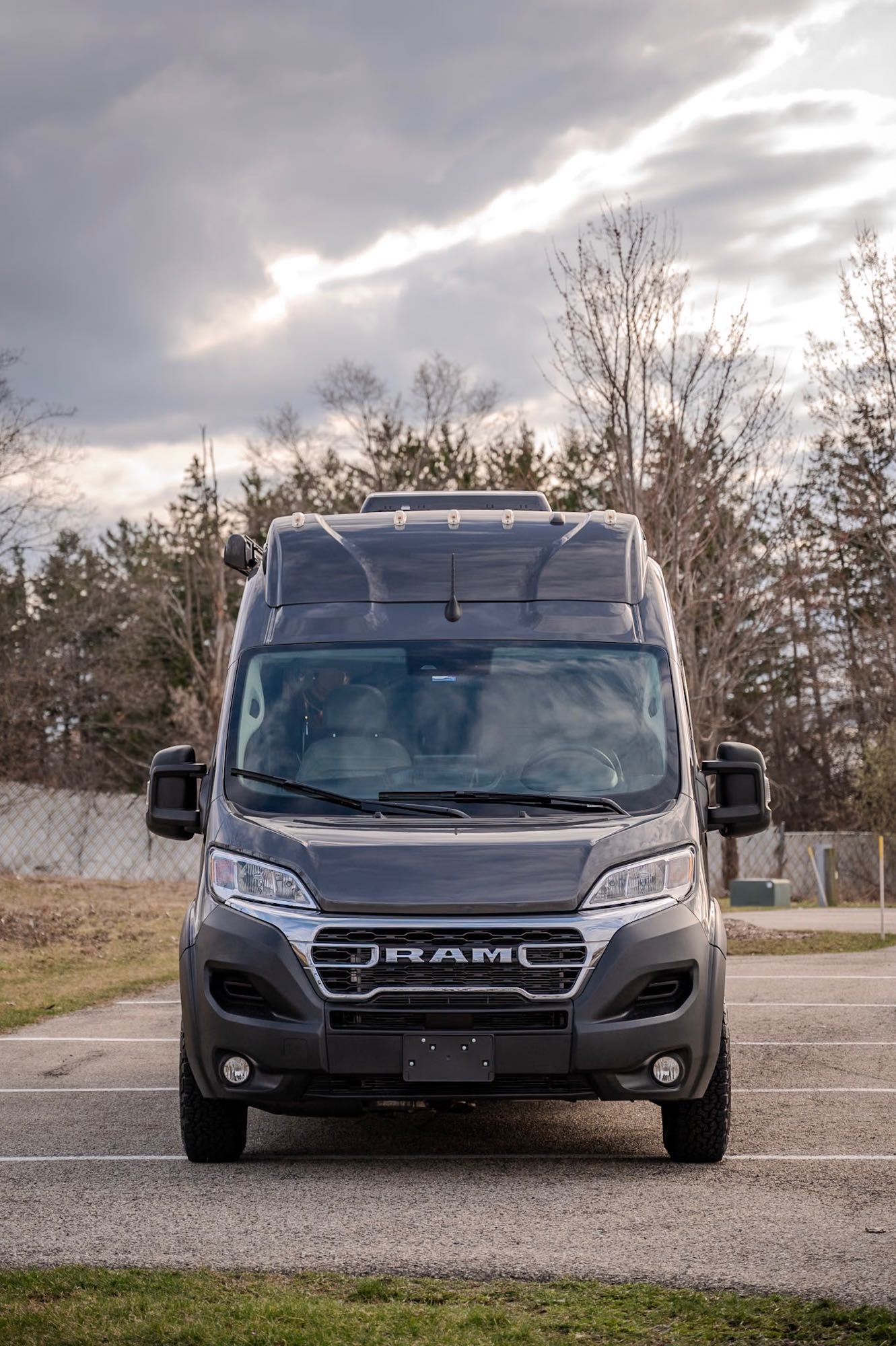 Dark gray Ram ProMaster camper van, a potential RV rental, seen head-on in a parking lot with trees.