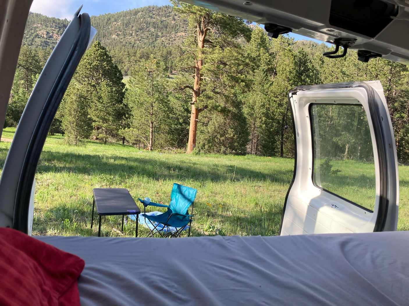 RV rental view from inside a camper van showing a serene outdoor camp setup with a table, chair, pine trees, and mountains.