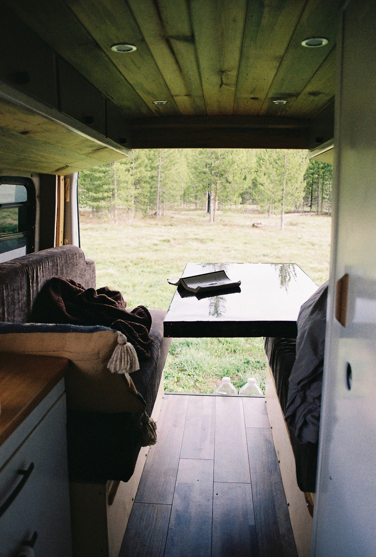 Inside a spacious rv rental. A book rests on a table between benches, with a clear view of a serene forest outside.
