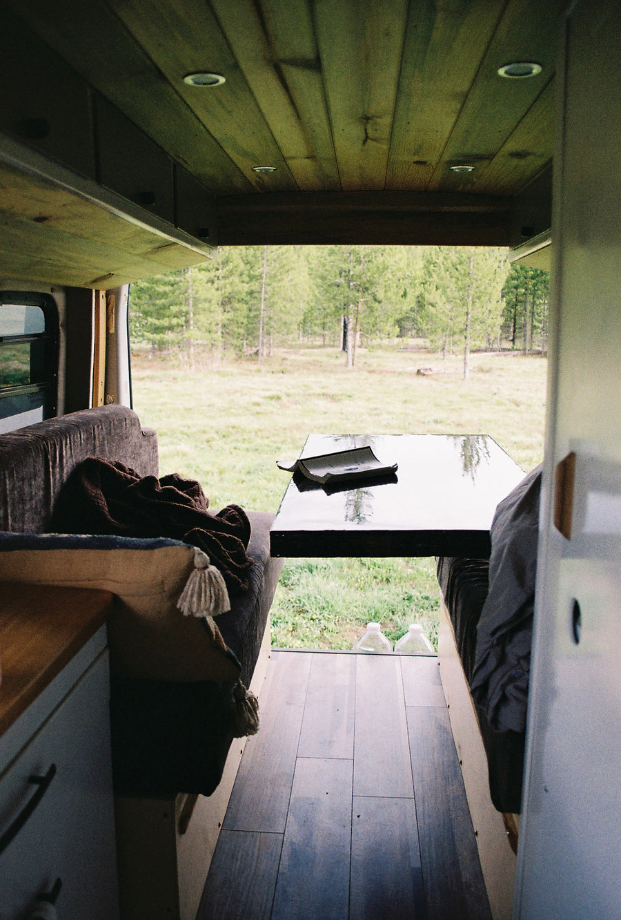 Inside a spacious rv rental. A book rests on a table between benches, with a clear view of a serene forest outside.