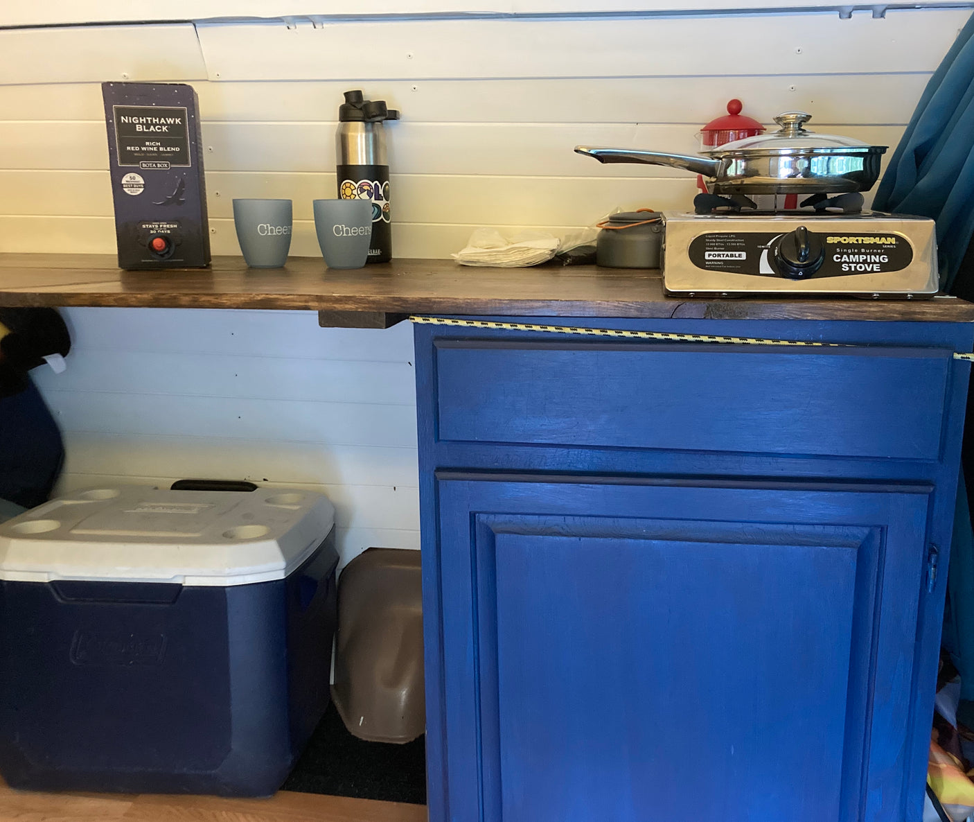 RV rental kitchenette features a portable camping stove, Nighthawk Black wine, and cooler, ideal for road trip cooking.