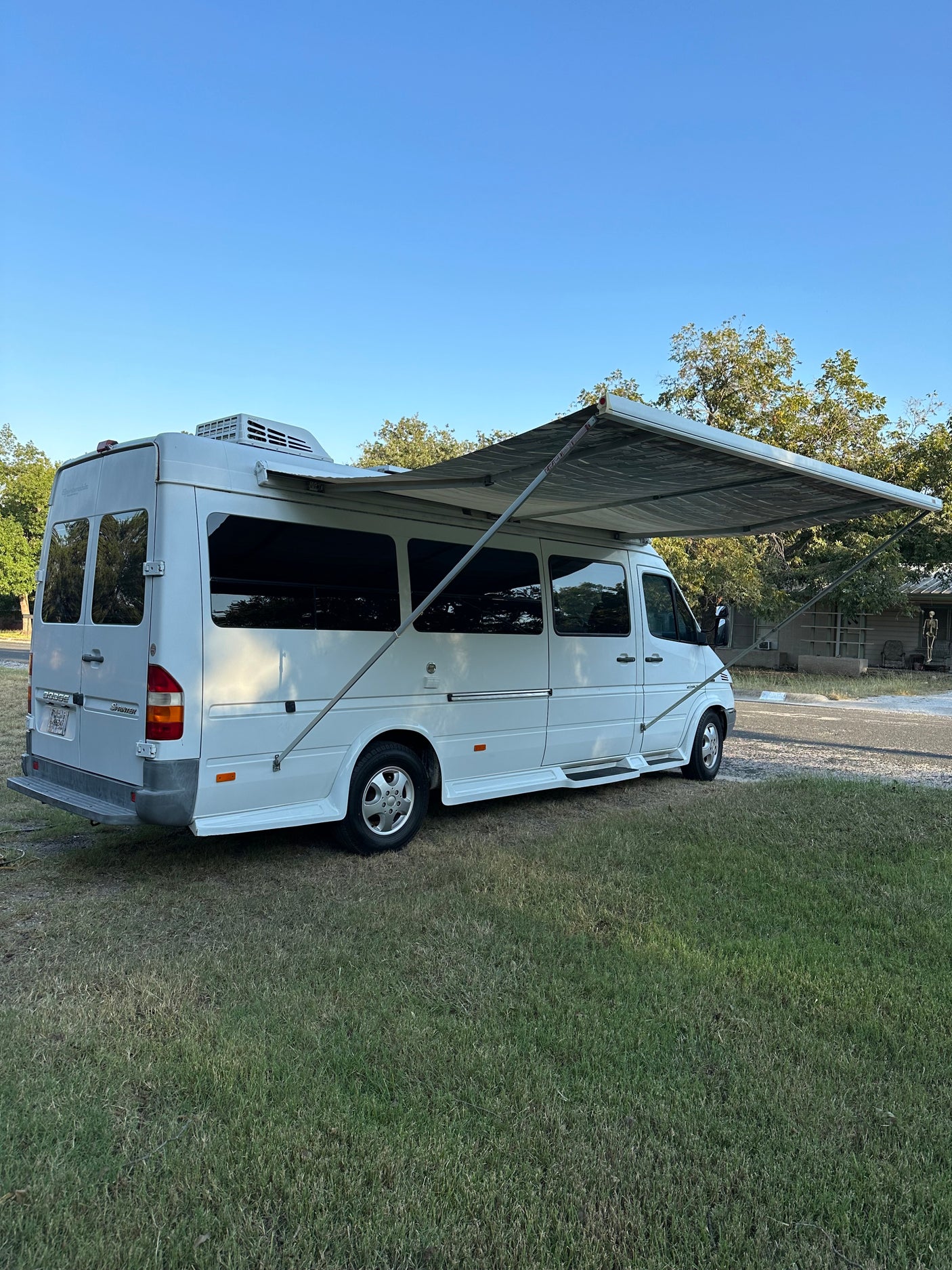 White Dodge Sprinter camper van with extended awning on grass, ready for an RV rental trip. Blue sky & trees in background.