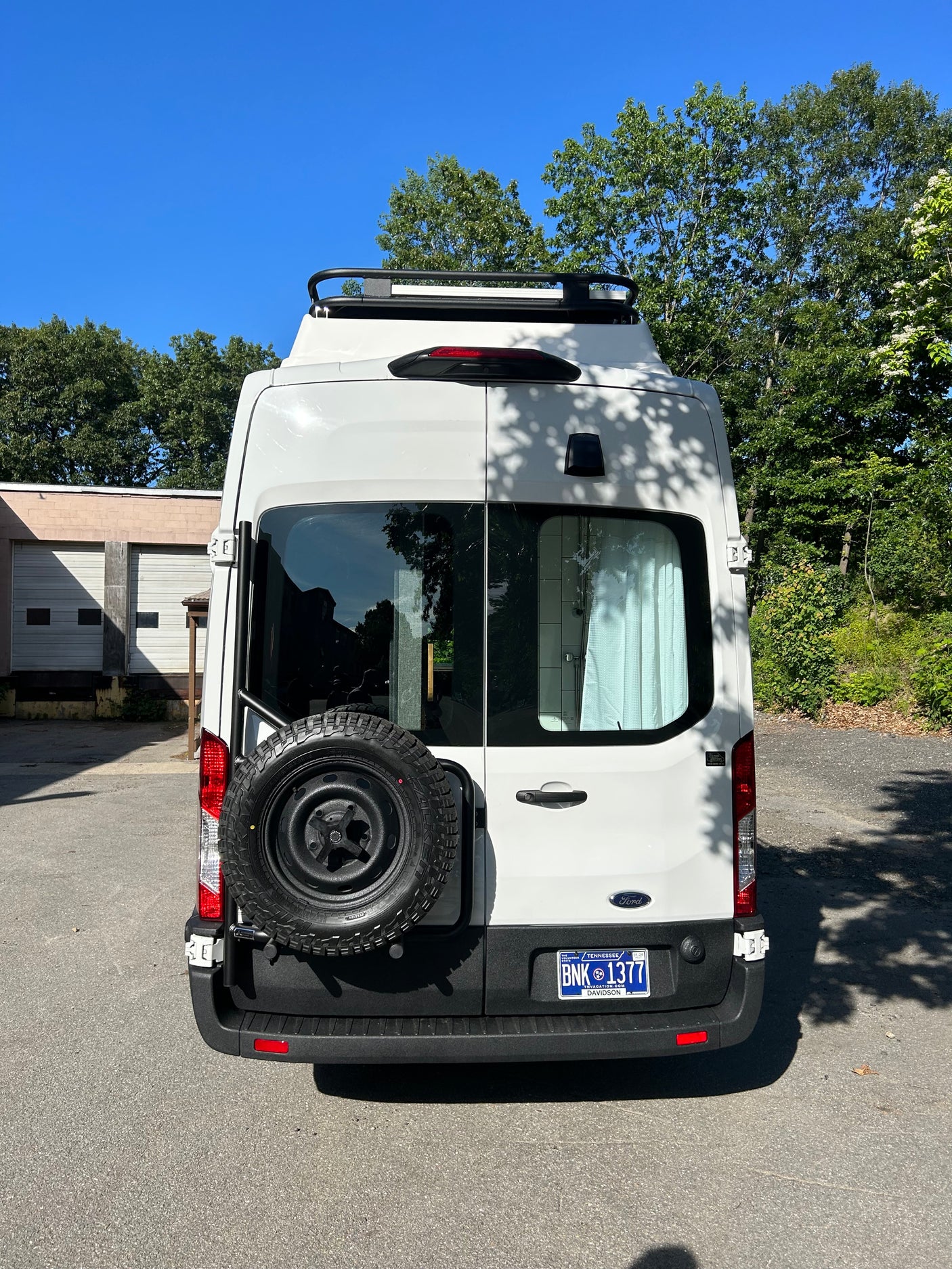 A white camper van for RV rental, seen from the rear with a spare tire mount and roof rack. Ready for travel adventures.