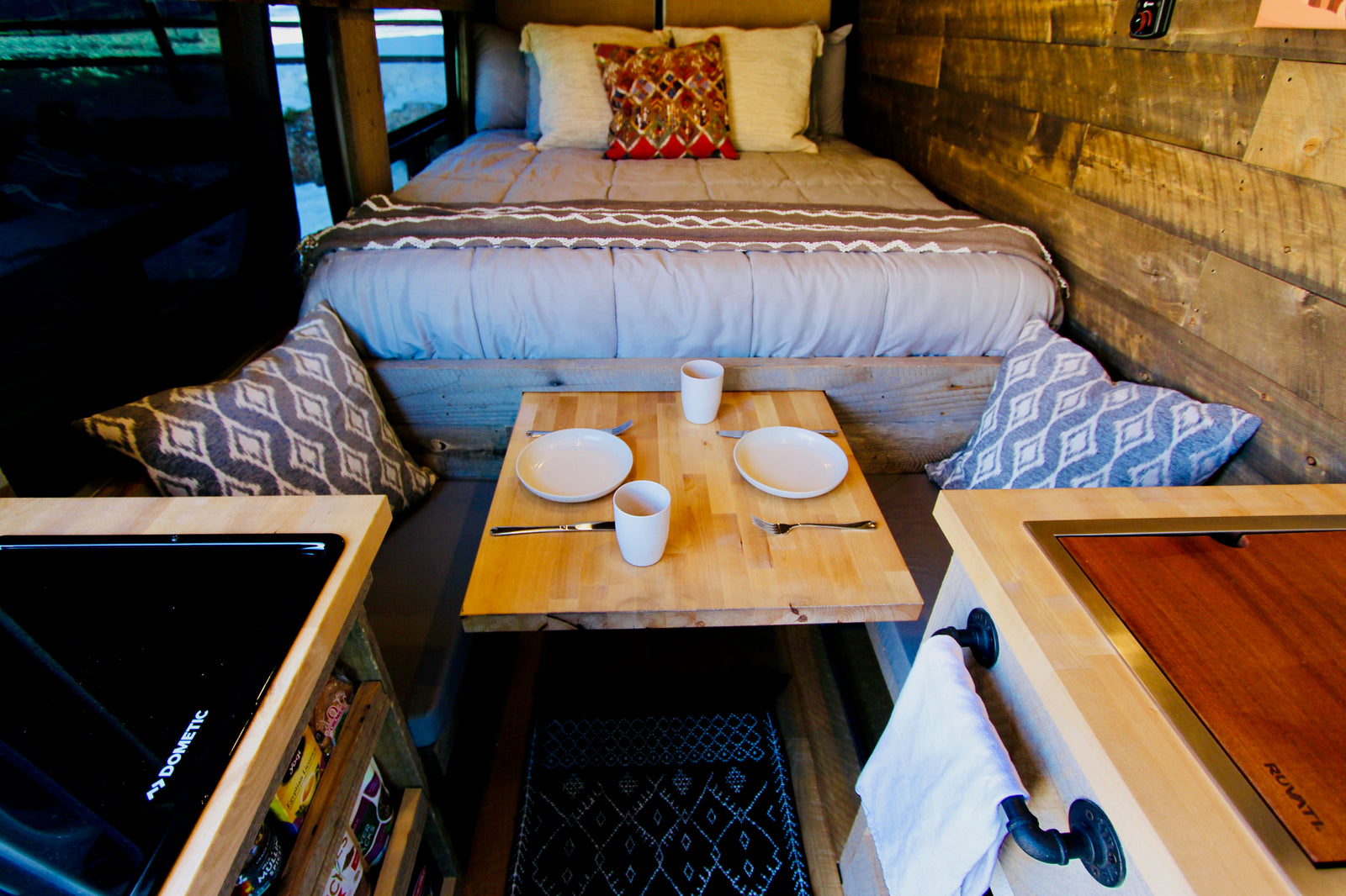 Cozy RV rental interior with a bed, dining table for two, Dometic stovetop, and Ruvati sink.
