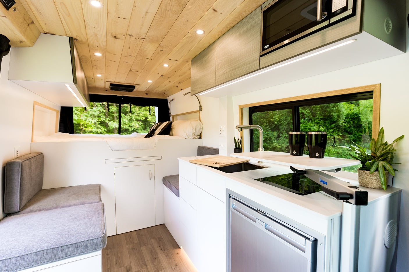 This stylish rv rental interior features a comfy bed, modern kitchen with fridge/cooktop, and a large window overlooking lush greenery.