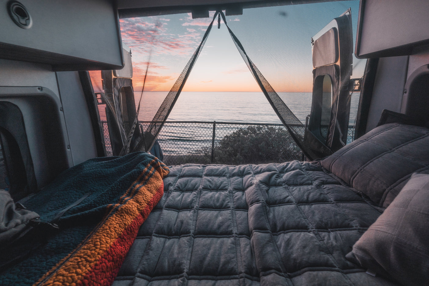 Cozy RV rental camper van bed facing an ocean sunset through screen-covered open doors, perfect for scenic retreats.
