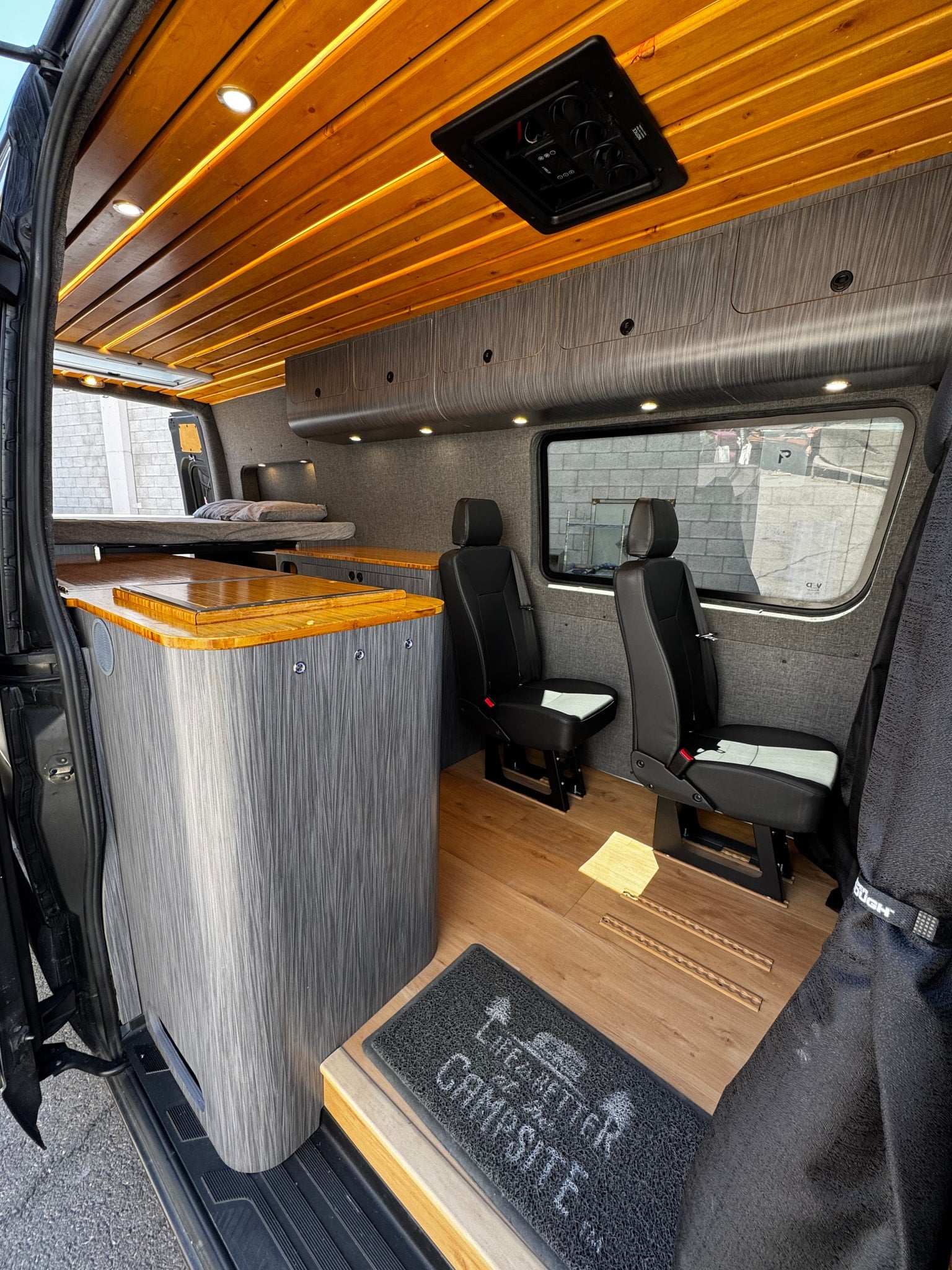 Modern RV rental interior: wood slat ceiling, gray cabinets, bed, kitchen counter, & passenger seats. Ready for adventure.